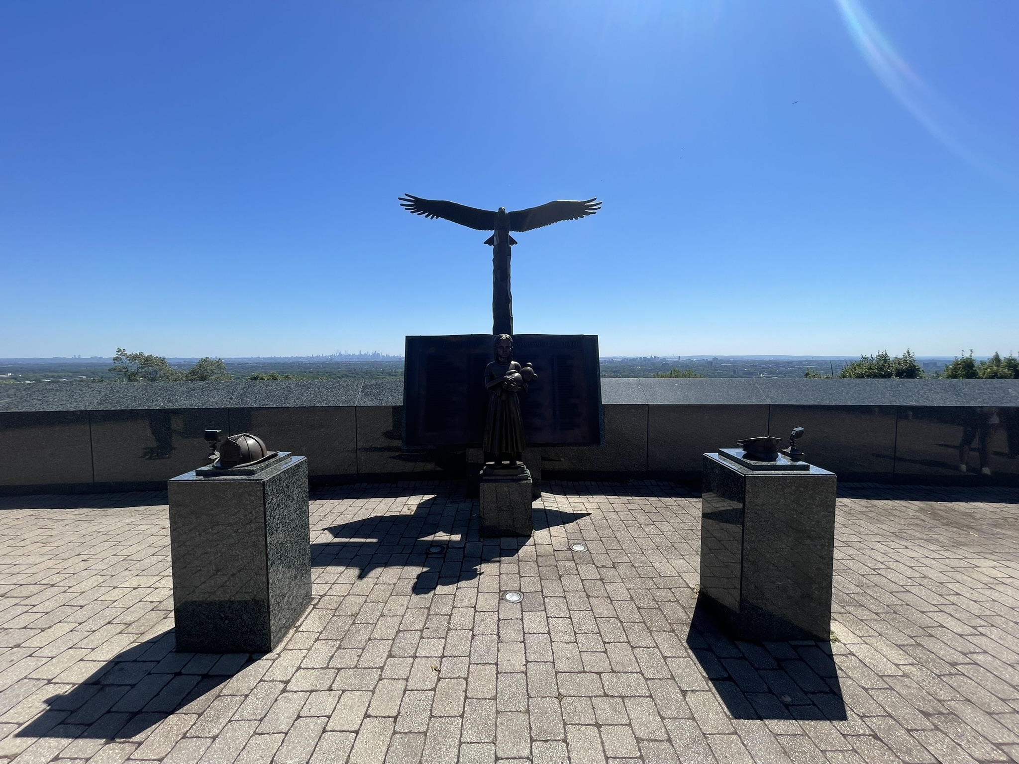 9/11 memorial at Eagle Rock Reservation featuring a bronze eagle with outstretched wings on a pedestal, flanked by memorial displays including a firefighter's helmet and police cap, with the New York City skyline visible in the distance